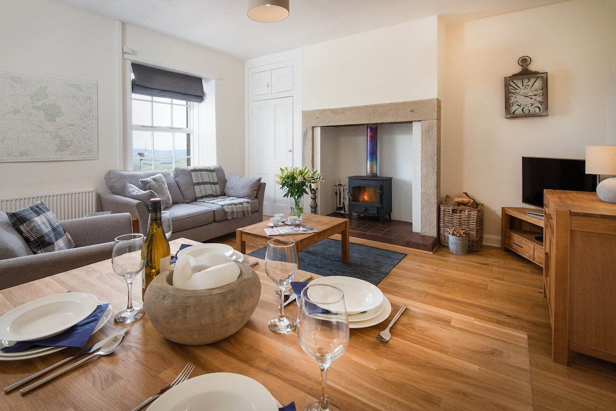 Chaffinch Cottage - sitting room with sofa, armchair, wood burning stove and dining area seating four guests